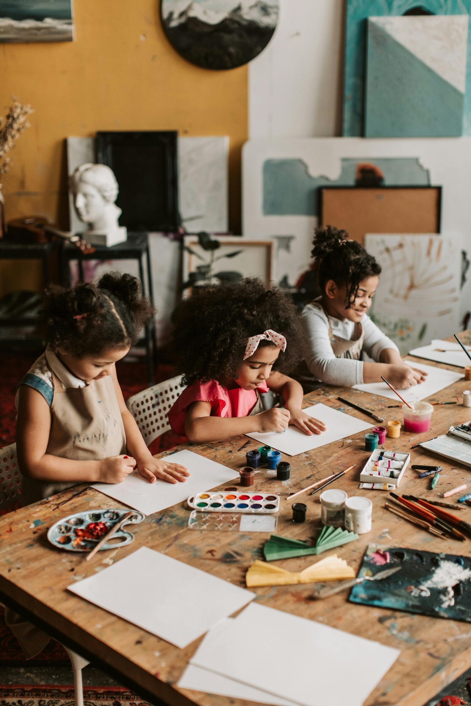 Three young girls engaged in painting at an indoor art workshop, using watercolors and brushes.